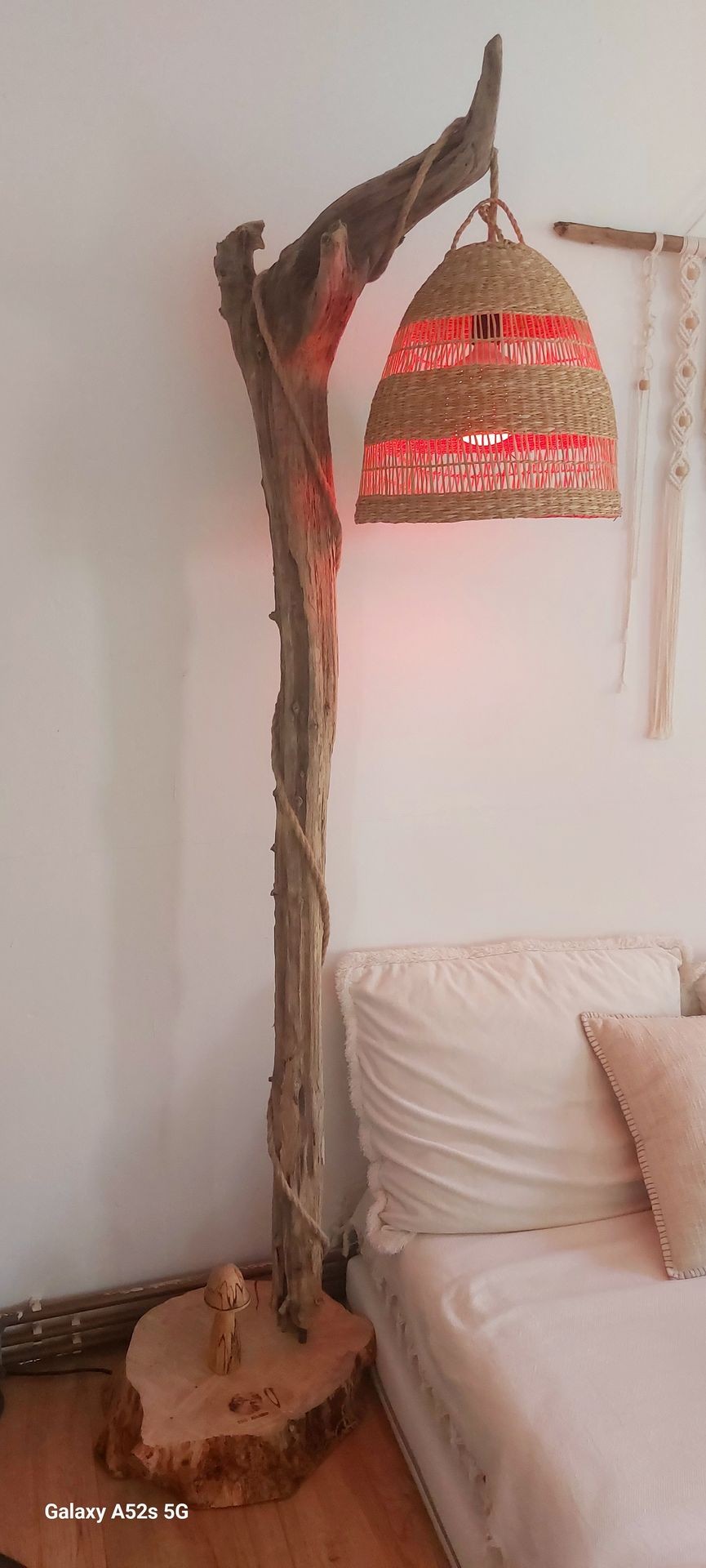 Wooden floor lamp with woven shade, emitting warm light next to a bed with white pillows.