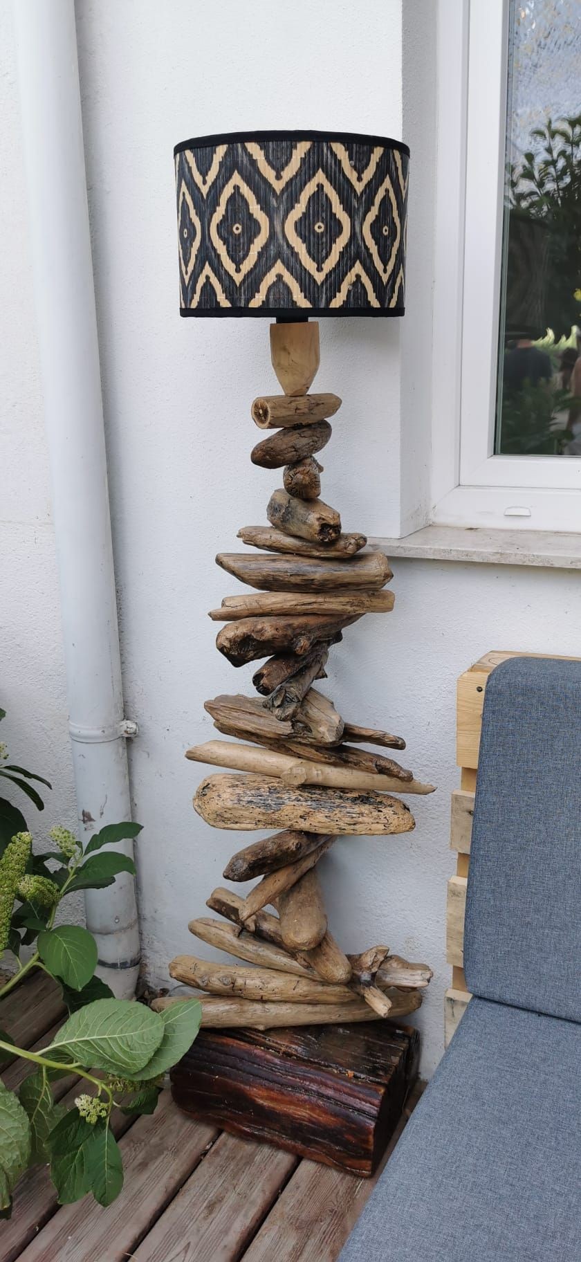 Driftwood floor lamp with patterned shade beside a window and grey seat on wooden deck.